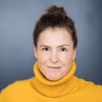 Kristina Dörlitz Frau mit lächelndem Gesicht und einem gelben Rollkragenpullover vor einem blauen Hintergrund.